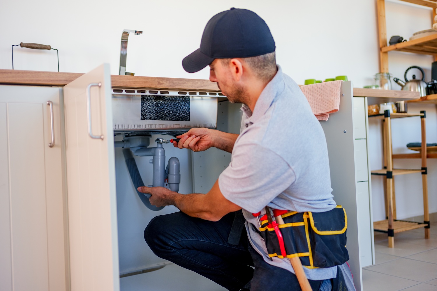 A plumber wearing a cap and tool belt repairs pipes under a kitchen sink, using a screwdriver while kneeling in front of an open cabinet.