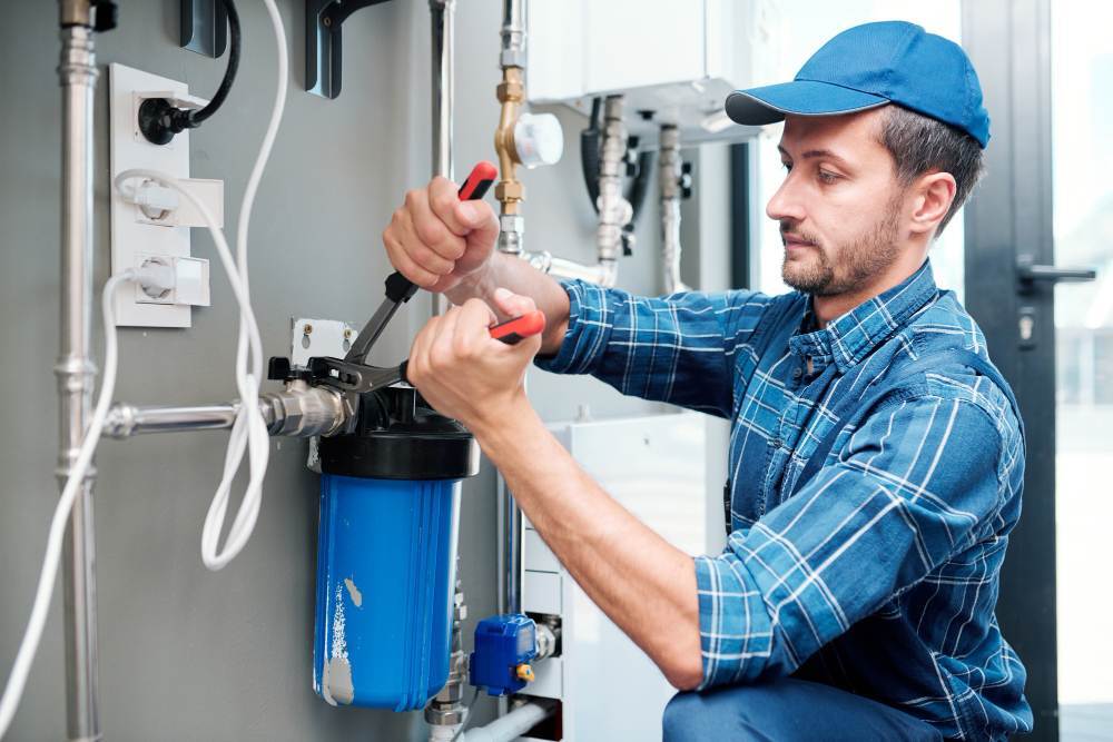 A plumber in a blue cap and plaid shirt uses tools to install or repair a water filtration system attached to a wall with pipes and wires.