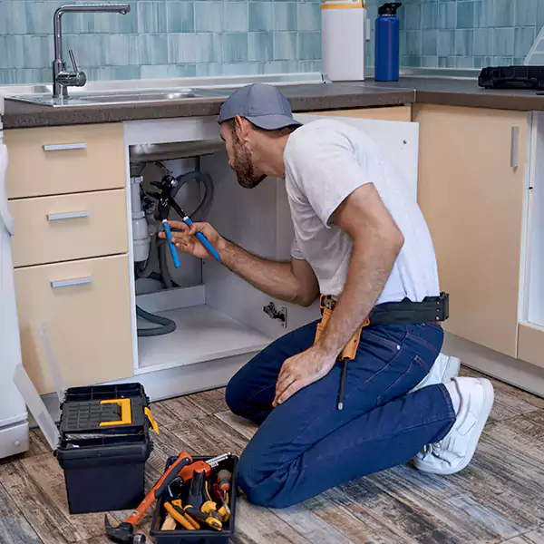 A plumber in a cap and work belt kneels on a kitchen floor, using a wrench to fix pipes under the sink. An open toolbox with various tools is beside him.