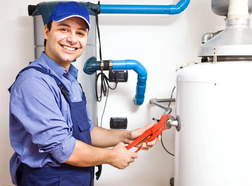 A smiling plumber in blue overalls and a cap uses a red pipe wrench to adjust a fitting on a water heater in a utility room with visible blue pipes.