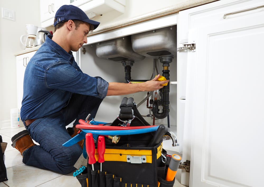 A plumber wearing a cap and denim clothes kneels on a kitchen floor, working under a sink. A tool bag filled with various tools sits nearby, and the cabinet doors are open to access the plumbing.
