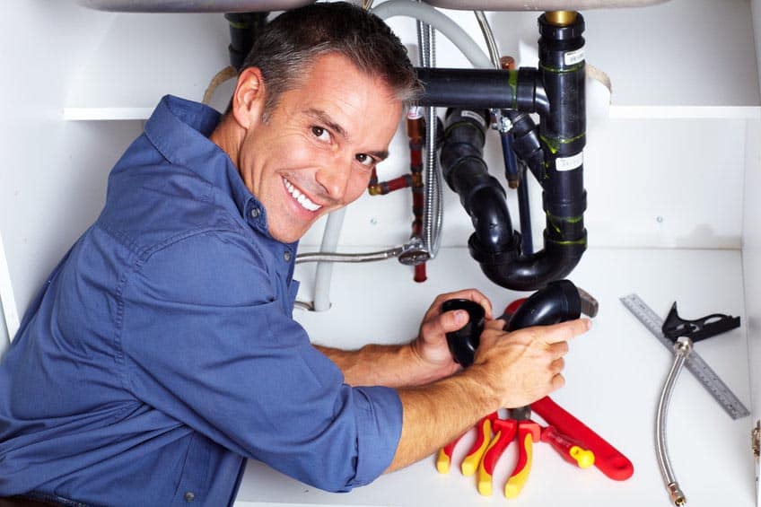 A smiling man in a blue shirt works under a sink, holding plumbing pipes and tools. He appears to be repairing or installing plumbing, surrounded by wrenches and pipe fittings.