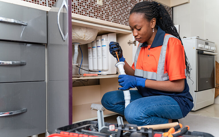 A woman in a uniform and gloves kneels on the floor, using a screwdriver to fix plumbing under a kitchen sink. A toolbox with various tools is open in front of her.