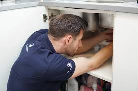 A person wearing a navy blue shirt is crouched under a kitchen sink, working on the plumbing inside a cabinet. Various cleaning supplies are visible in the cabinet below.